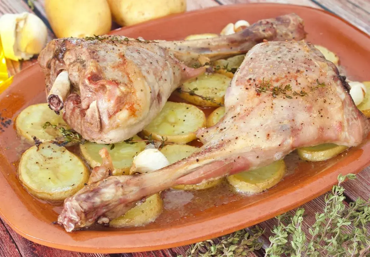 Dos jugosas piernas de cabrito al horno, tiernas y doradas, reposan sobre patatas asadas y dientes de ajo.