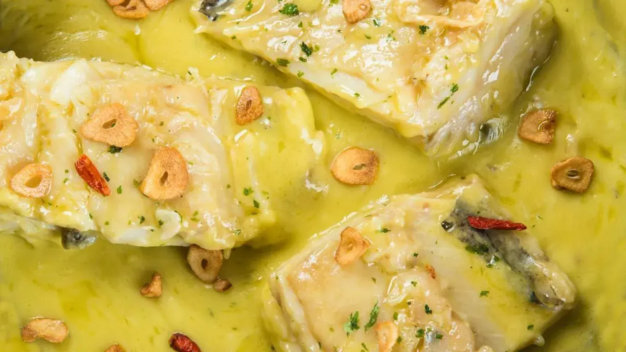 Merluza al pil pil, un plato tradicional con trozos de pescado blanco en una salsa cremosa, adornado con láminas de ajo dorado y guindilla.