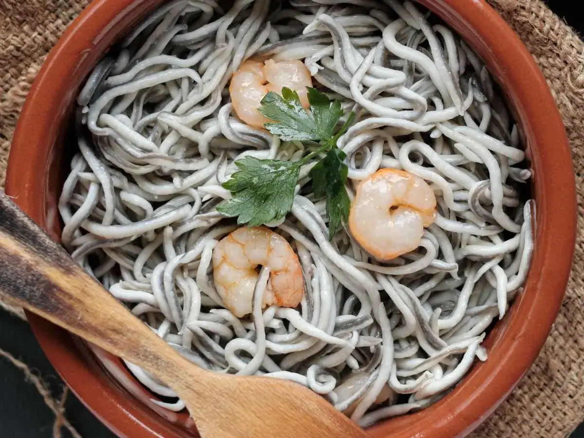 Gulas al ajillo servidas en cazuela de barro, con gambas y perejil. Una cuchara de madera lista para servir.