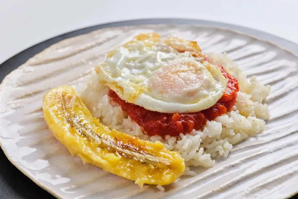 Un plato de arroz a la cubana con huevo frito, salsa roja y plátano maduro frito.