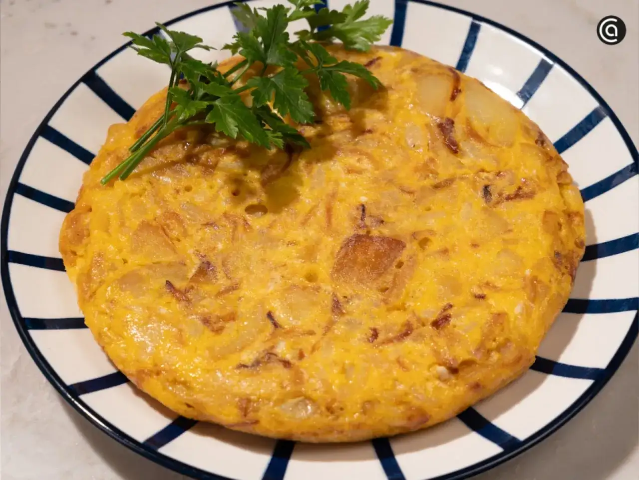 Tortilla de patatas dorada y esponjosa, decorada con perejil fresco, servida en un plato a rayas azules.