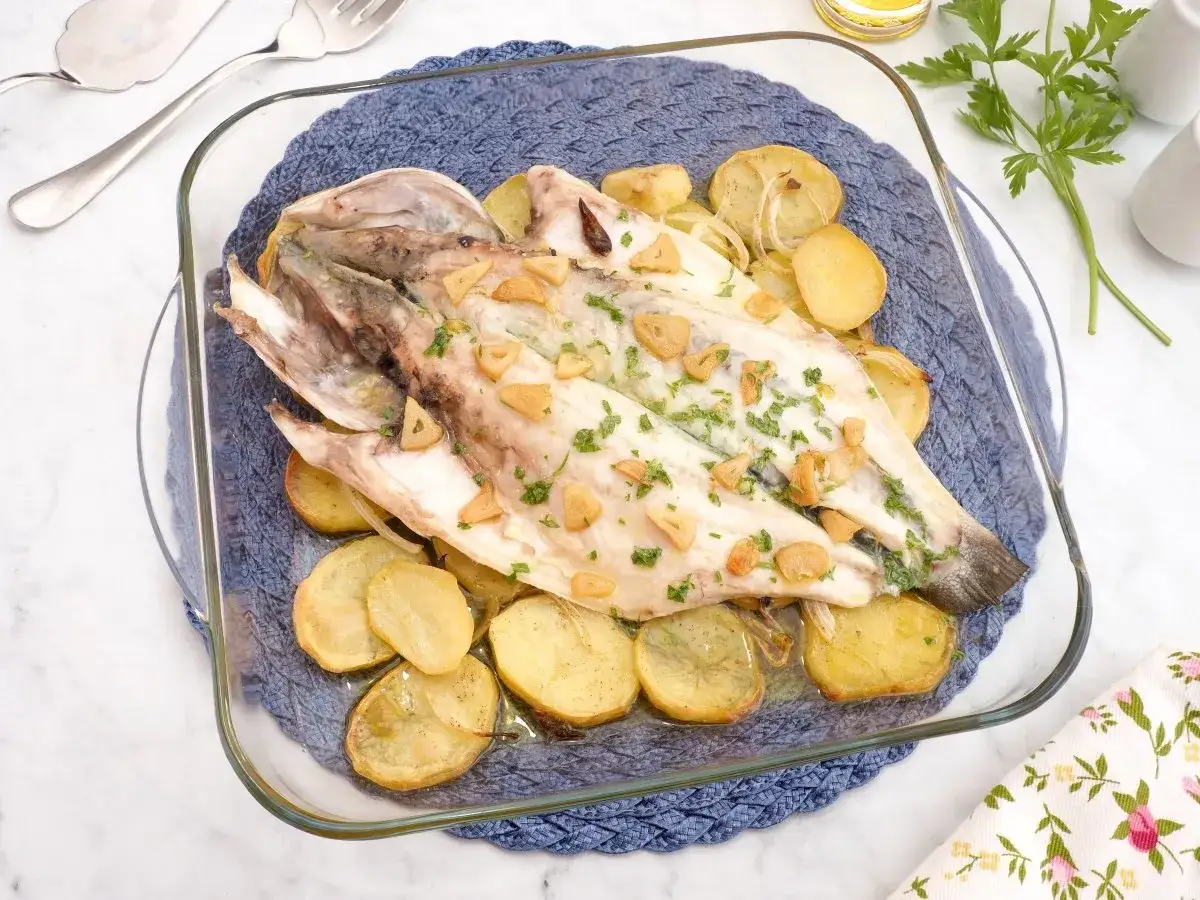 Lubina a la espalda horneada con patatas y ajo, servida en una fuente de cristal sobre un salvamanteles azul.