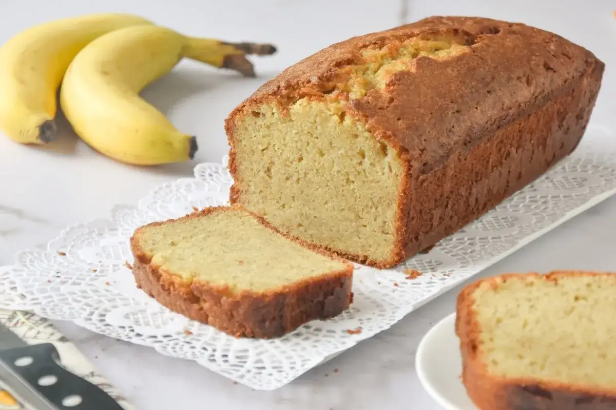 Un delicioso pastel de pl&aacute;tano reci&eacute;n horneado, con una rebanada cortada y dos pl&aacute;tanos amarillos al fondo.