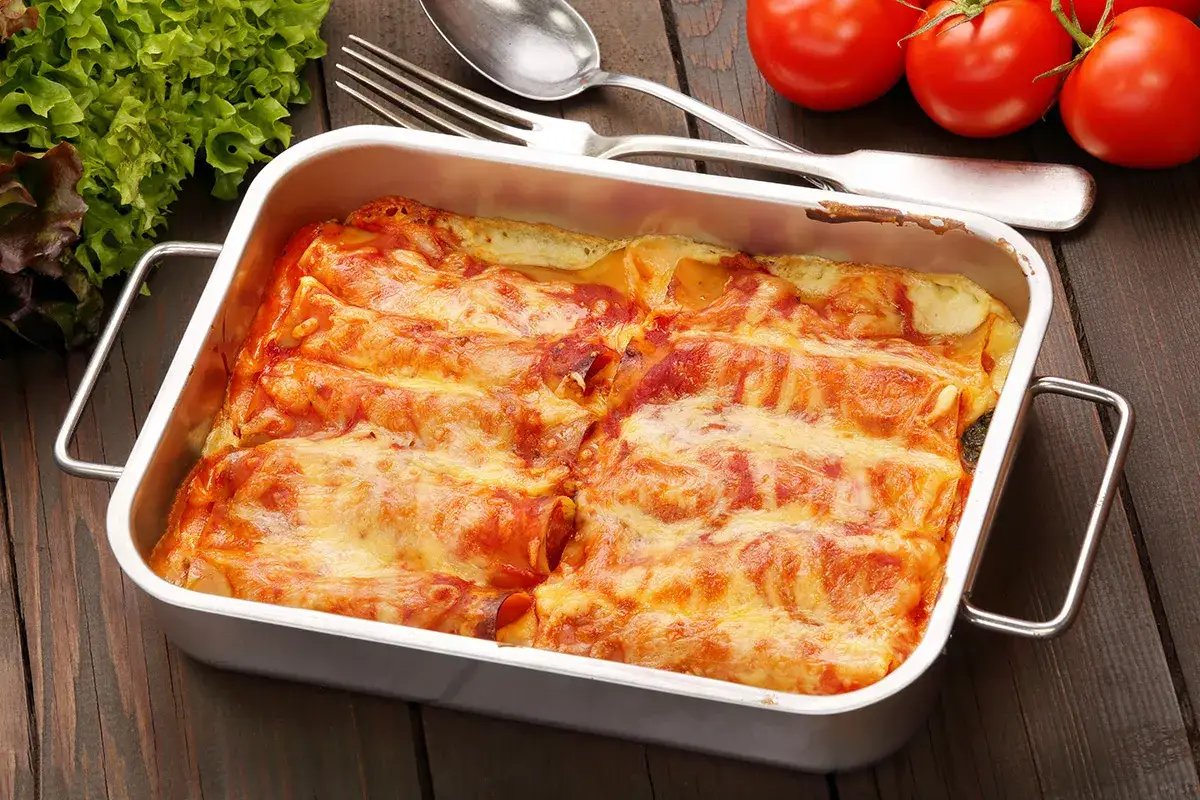 Canelones de at&uacute;n gratinados en una bandeja met&aacute;lica, acompa&ntilde;ados de lechuga fresca y tomates.