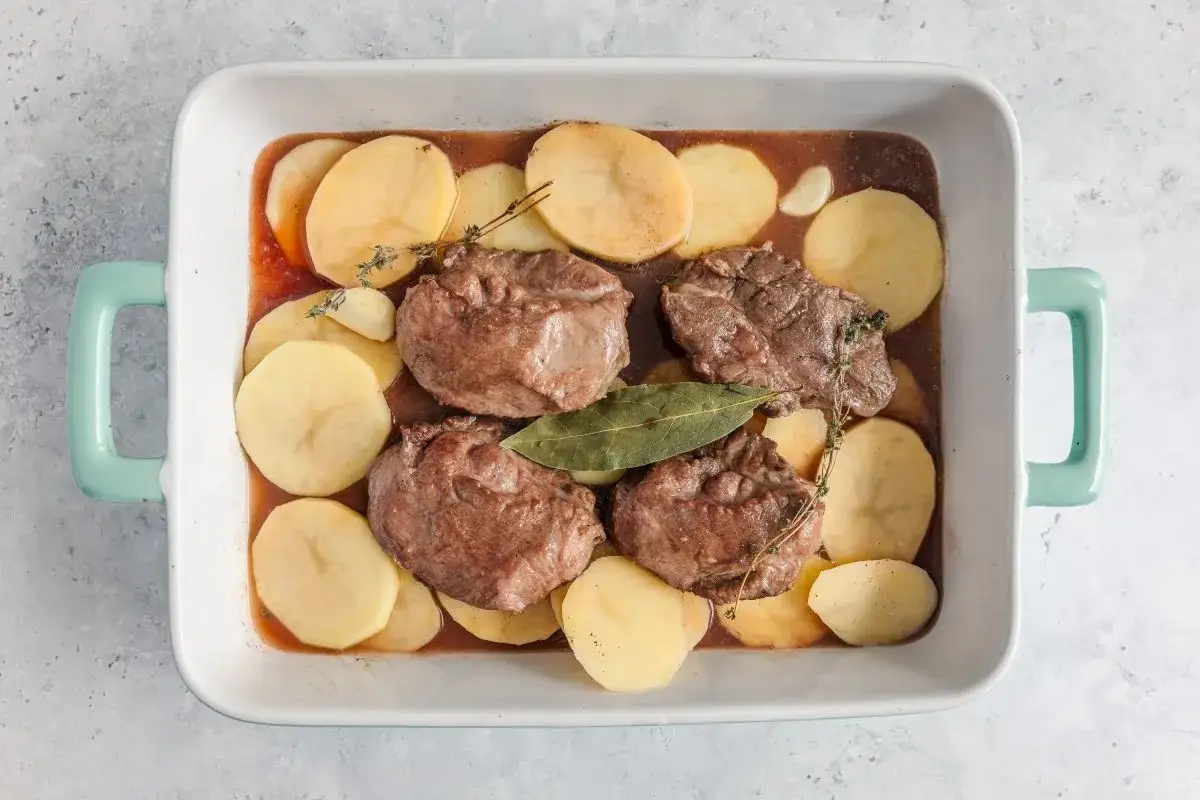 Cuatro trozos de carne tierna y patatas en rodajas en una fuente para horno, listas para unas deliciosas galtas al horno.