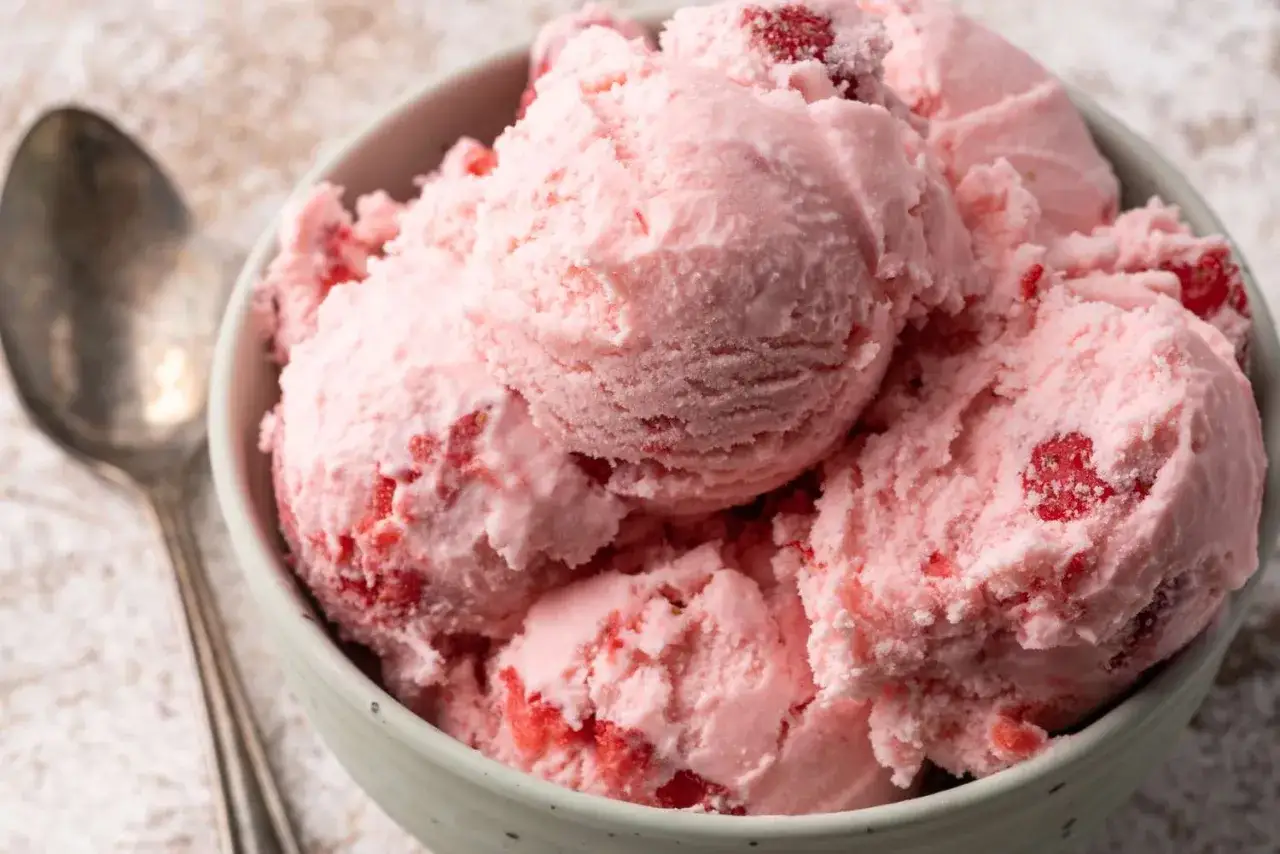 Un taz&oacute;n lleno de cremoso helado de fresa, con trozos de fruta roja visible. Una cuchara plateada descansa a un lado.