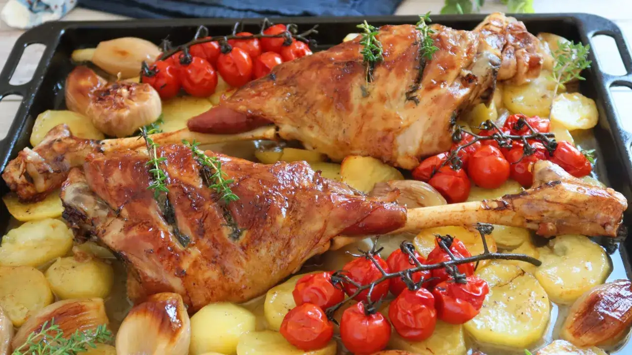 Dos jugosas piernas de cordero al horno, doradas y tiernas, reposan sobre patatas asadas, tomates cherry y cebollas.