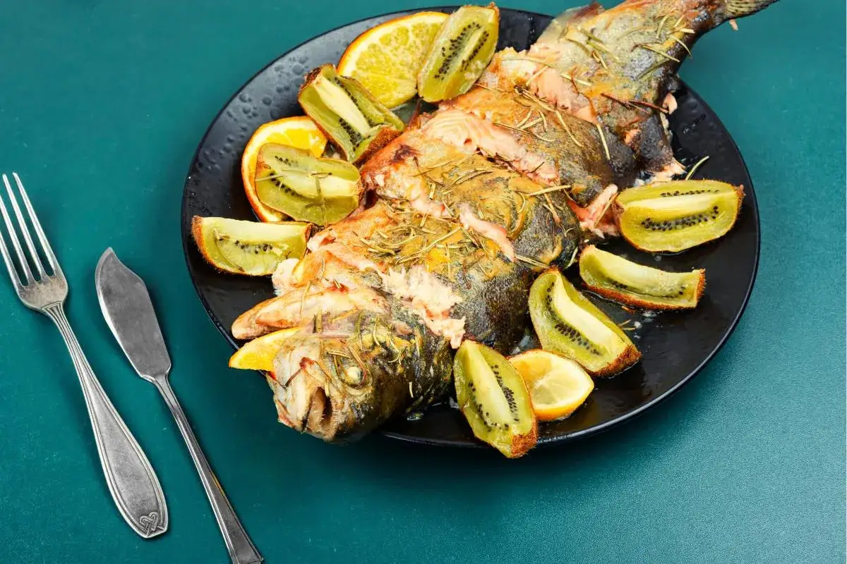 Trucha al horno con rodajas de naranja y kiwi, servida en un plato negro con cubiertos.