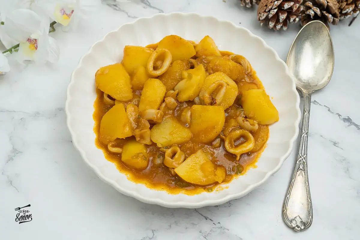 Plato de patatas con calamares en salsa, servido en un plato blanco con un borde ondulado. Una cuchara plateada reposa a la derecha.