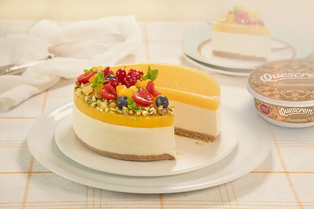 Tarta de mango con frutas rojas y pistachos. Una porci&oacute;n cortada revela su interior cremoso.