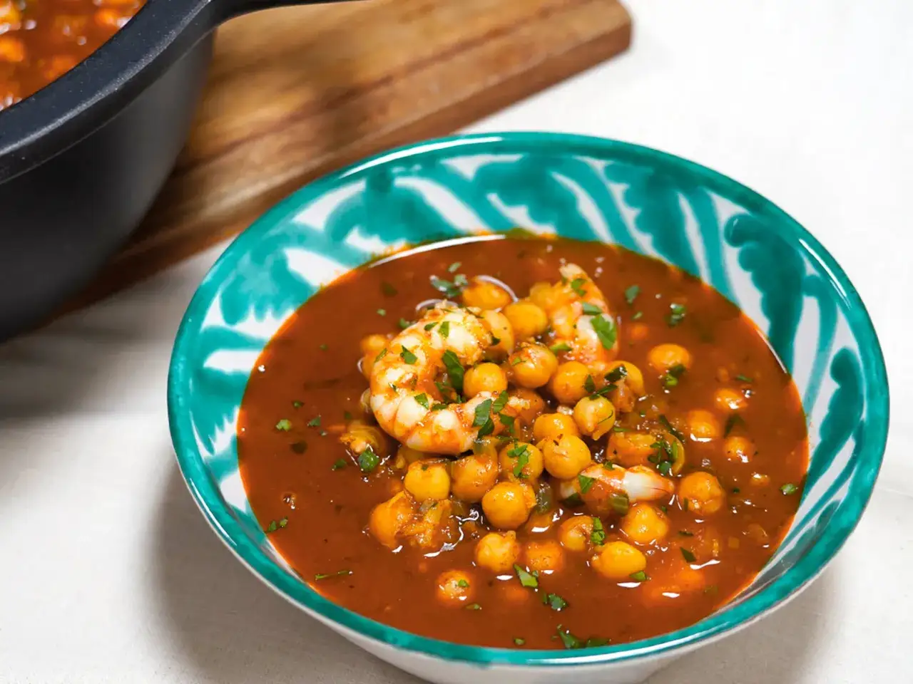 Un taz&oacute;n de garbanzos con langostinos en una salsa rojiza, adornado con perejil picado.