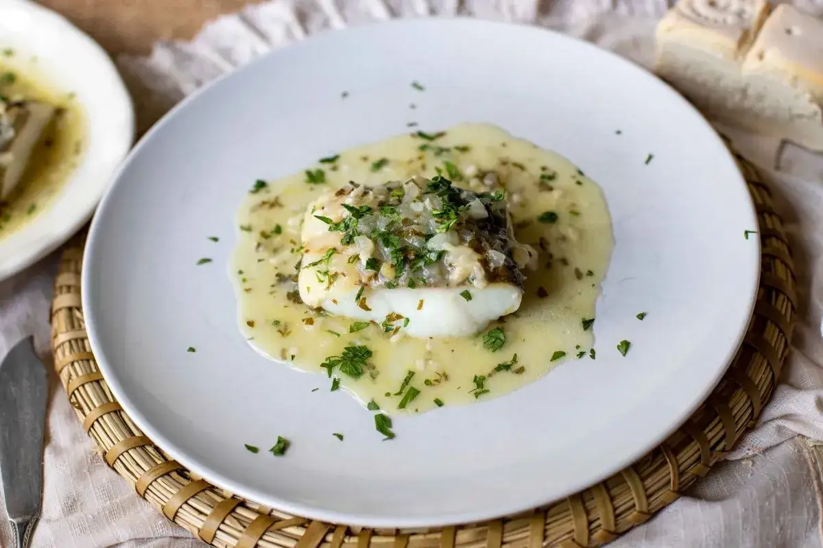 Delicioso bacalao en salsa verde, con trocitos de cebolla y perejil fresco, servido en un plato blanco.