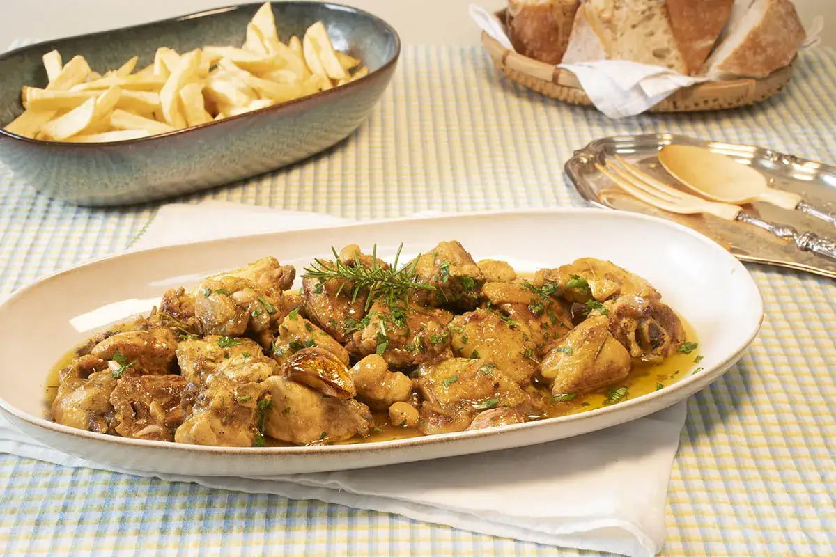 Sabrosos muslos de pollo guisados con hierbas y ajo, servidos en una fuente ovalada. Acompa&ntilde;ados de patatas fritas y pan.