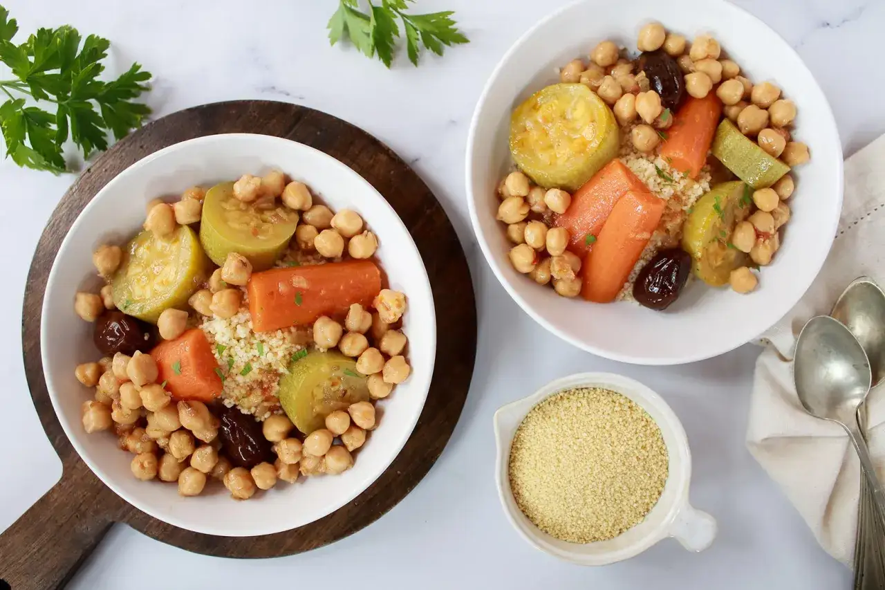 Dos cuencos de cous cous receta con garbanzos, zanahoria, calabac&iacute;n y ciruelas pasas. Un cuenco de s&eacute;mola de trigo al lado.
