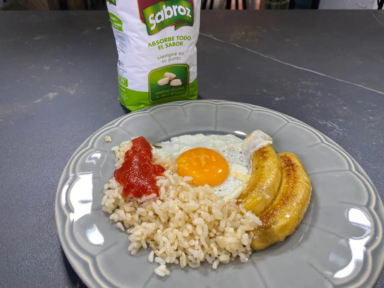 Plato de arroz a la cubana con huevo frito, plátanos fritos y salsa de tomate. Bolsa de arroz Sabroz al fondo.