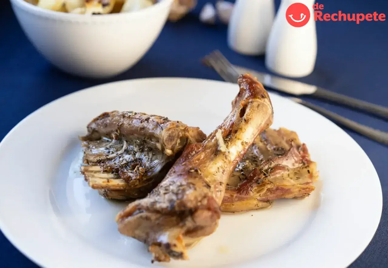 Delicioso cabrito al horno, tierno y dorado, servido en un plato blanco. Acompa&ntilde;ado de patatas asadas en un cuenco.