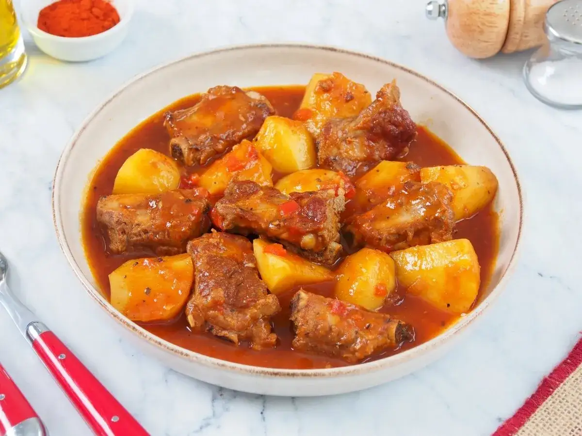 Plato humeante de patatas con costillas en salsa roja, con trozos de pimiento rojo y especias.