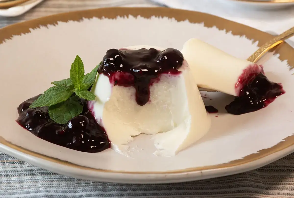 Deliciosa panna cotta receta con salsa de frutos rojos y menta fresca en un plato blanco con borde dorado.