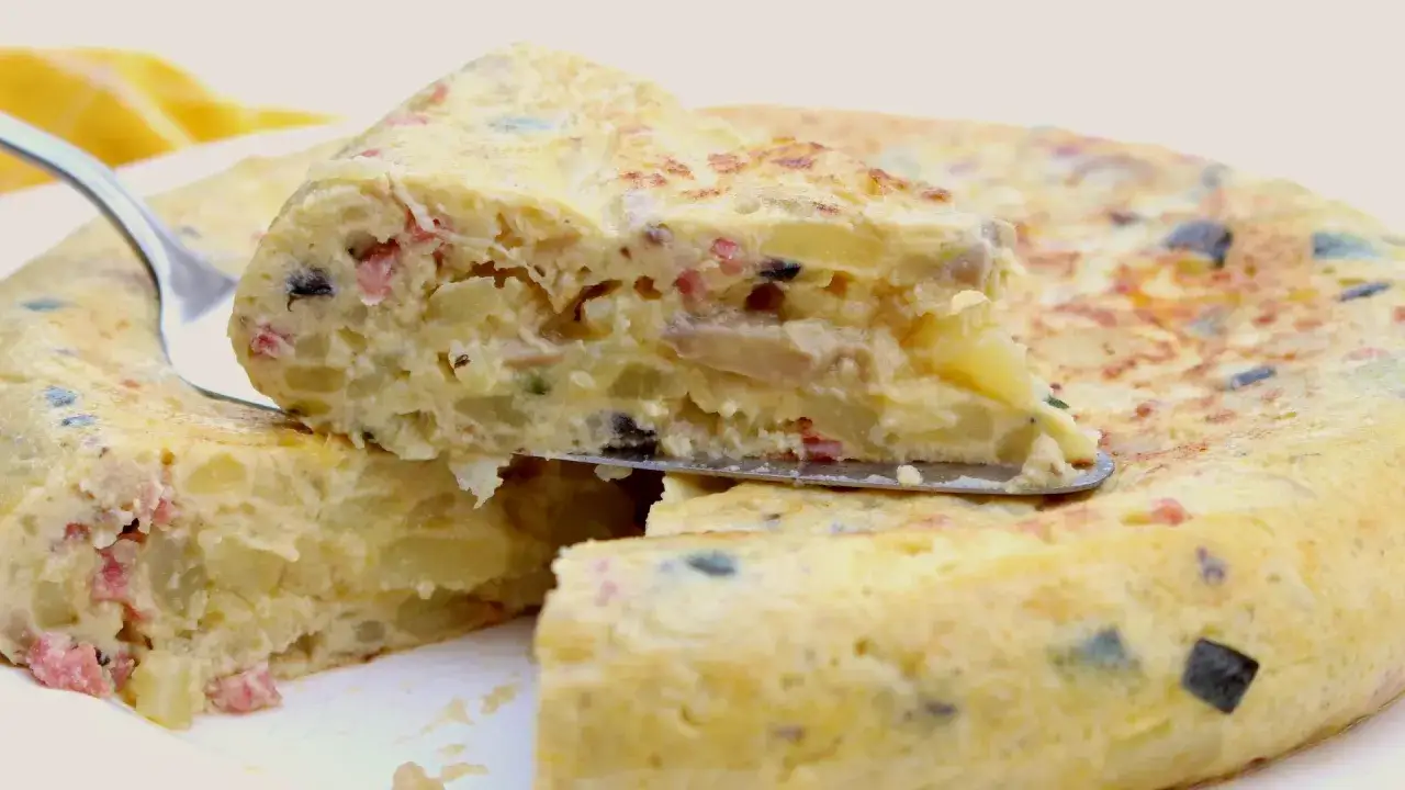 Una porción de tortilla de patatas, jugosa y con trozos de jamón y aceitunas, se levanta con una pala.