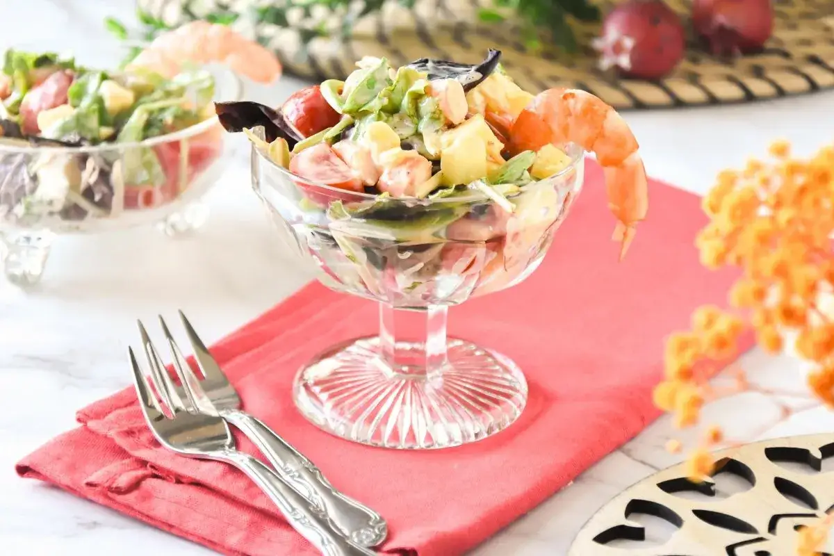 Delicioso coctel de marisco con gambas, tomates y lechuga en copa de cristal. Servido sobre mantel rosa con tenedores.