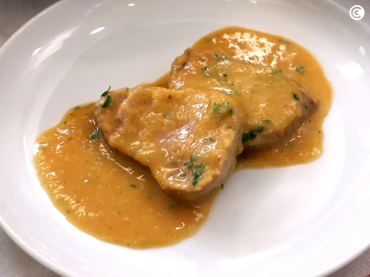Dos trozos de solomillo de cerdo en una cremosa salsa dorada con perejil picado, servidos en un plato blanco.