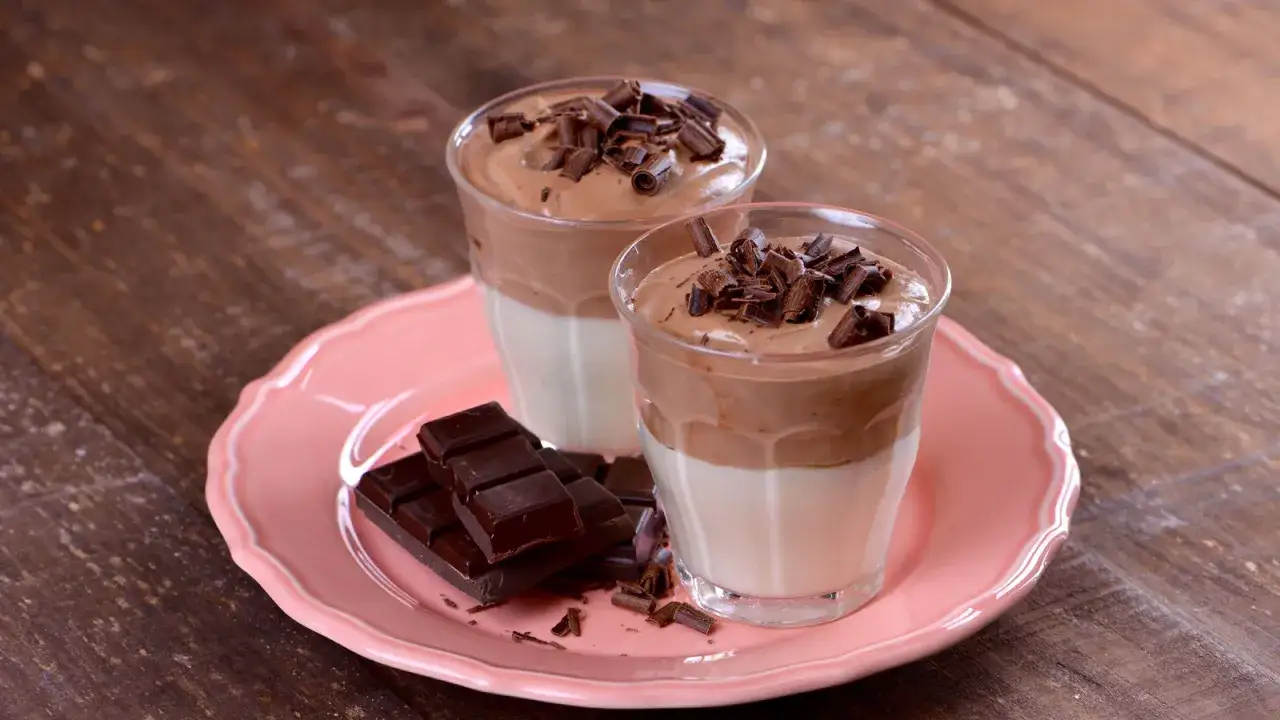Dos copas de mousse de chocolate con virutas de chocolate encima, junto a trozos de chocolate negro en un plato rosa.