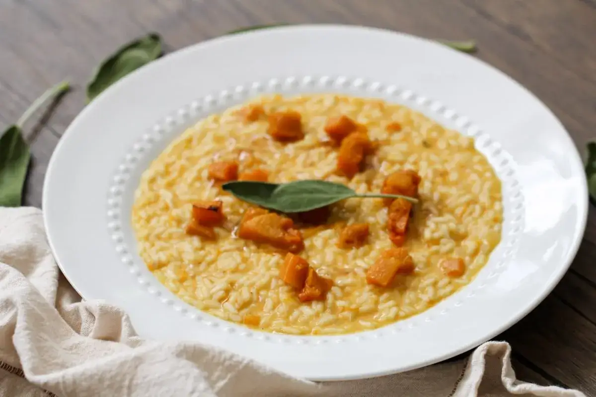 Un cremoso risotto de calabaza, adornado con trozos de calabaza asada y una hoja de salvia fresca, servido en un plato blanco.