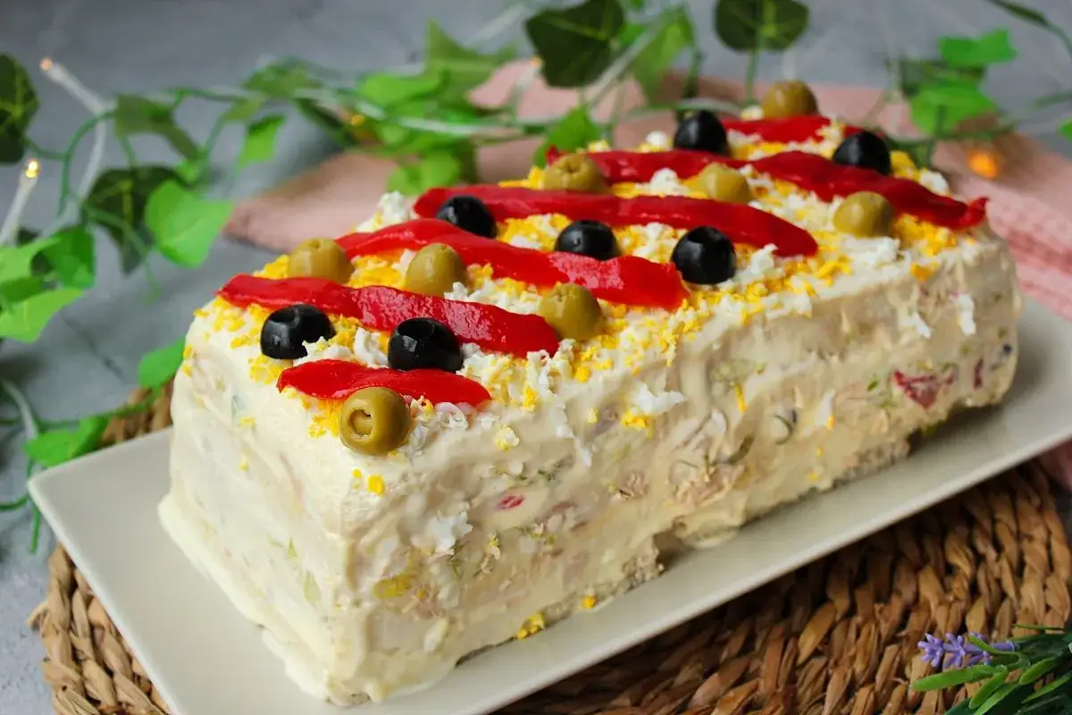 Pastel de at&uacute;n rectangular cubierto de mayonesa, decorado con pimientos rojos, aceitunas negras y verdes, y huevo rallado.