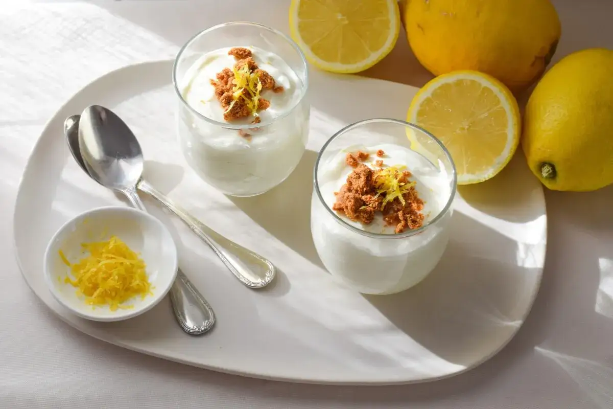 Dos copas de mousse de lim&oacute;n con crumble y ralladura, acompa&ntilde;adas de limones frescos y cucharas.