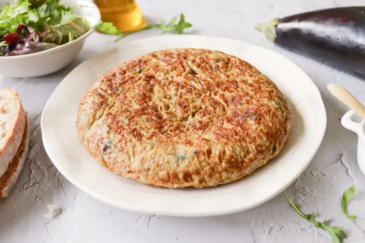 Tortilla de berenjena dorada y esponjosa, servida en plato blanco, acompa&ntilde;ada de pan y ensalada.