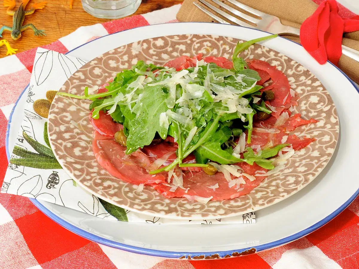 Delicioso carpaccio de ternera con r&uacute;cula fresca y queso parmesano rallado, servido en un plato decorado.
