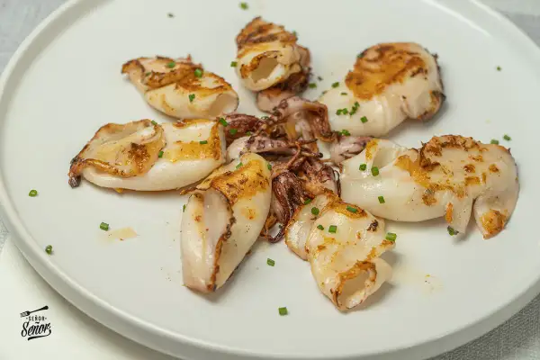 Deliciosos chipirones a la plancha, dorados y tiernos, servidos en un plato blanco con cebollino picado.