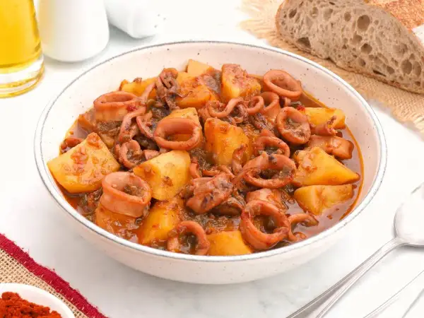 Un tazón de patatas con calamares en salsa, acompañado de pan rústico y una pizca de pimentón.
