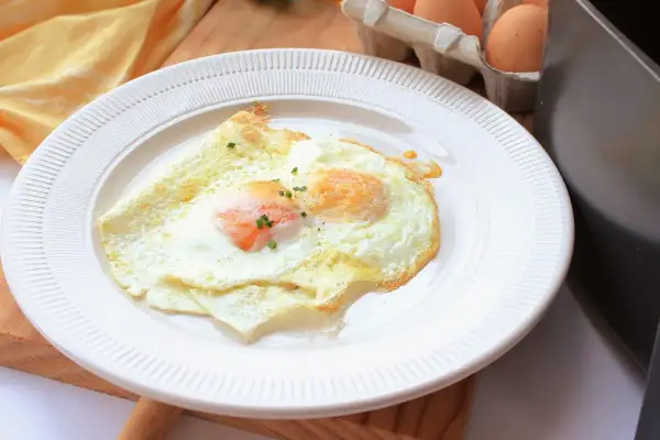Dos huevos fritos perfectos, cocinados en freidora de aire, servidos en un plato blanco. Un toque de cebollino fresco adorna las yemas.