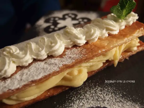 Delicioso milhojas de crema, con hojaldre crujiente y relleno cremoso, decorado con nata montada y una hoja de menta.