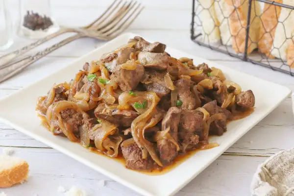 Sabrosos higaditos de pollo encebollados, servidos en un plato blanco, con pan y tenedores al fondo.