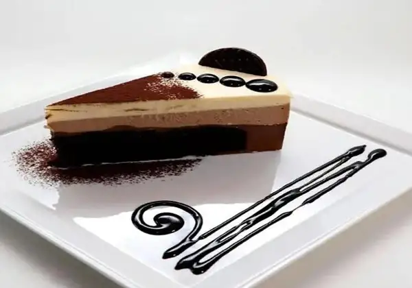 Deliciosa tarta de 3 chocolates, con capas de mousse y bizcocho, decorada con cacao y un toque de chocolate líquido.