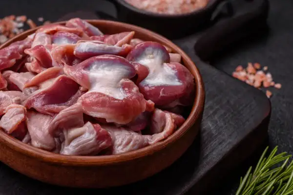 Un cuenco de madera lleno de mollejas de pollo crudas, listas para ser cocinadas. Sal rosada y romero al lado.