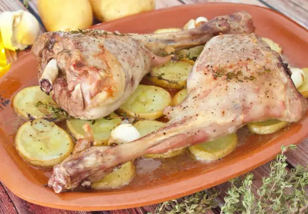 Dos jugosas piernas de cabrito al horno, tiernas y doradas, reposan sobre patatas asadas y dientes de ajo.