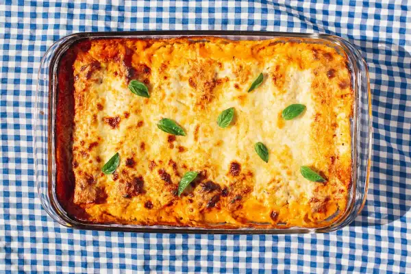 Bandeja de canelones de espinacas gratinados, decorados con hojas de albahaca fresca, sobre un mantel de cuadros azules y blancos.