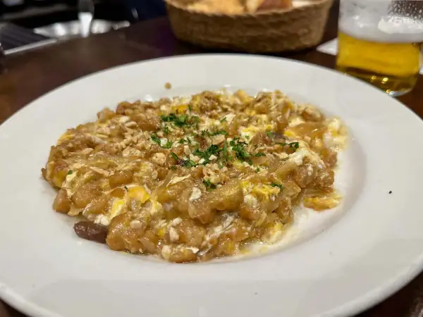 Delicioso revuelto de bacalao con trocitos de frutos secos y perejil picado, servido en plato blanco.
