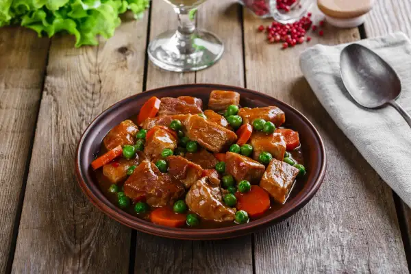 Un plato de carne en salsa con guisantes y zanahorias, servido en una cazuela de barro sobre una mesa de madera rústica.