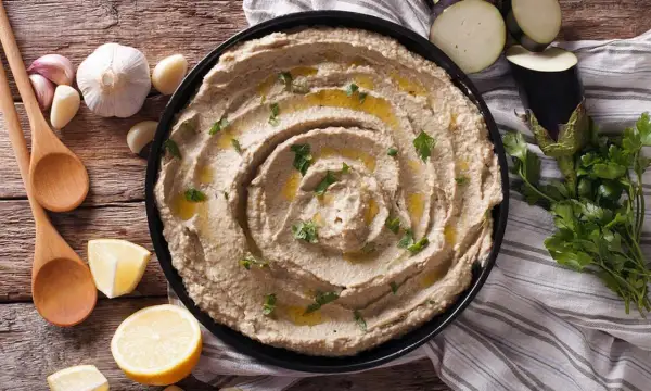 Un cremoso hummus de berenjena, adornado con aceite de oliva y perejil, junto a limones, ajos y cucharas de madera.