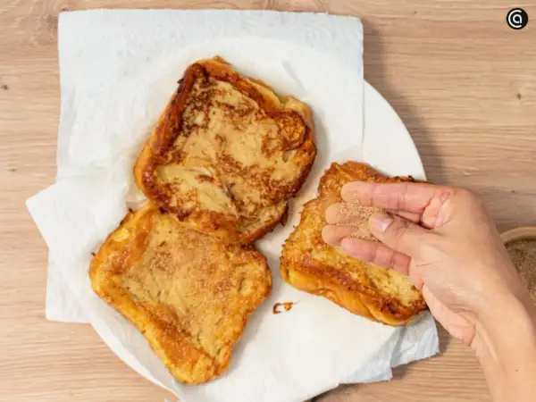 Torrijas de horchata doradas y crujientes, listas para disfrutar. Una mano espolvorea azúcar y canela sobre una de ellas.