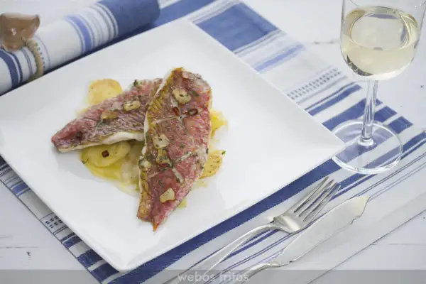 Dos deliciosos salmonetes al horno, con patatas y ajo, servidos en un plato blanco junto a una copa de vino blanco.