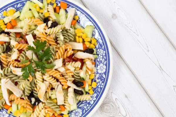 Ensalada de pasta con pollo, pepino, maíz y pasas, decorada con perejil fresco. Un plato colorido y nutritivo.