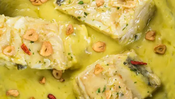 Merluza al pil pil, un plato tradicional con trozos de pescado blanco en una salsa cremosa, adornado con láminas de ajo dorado y guindilla.
