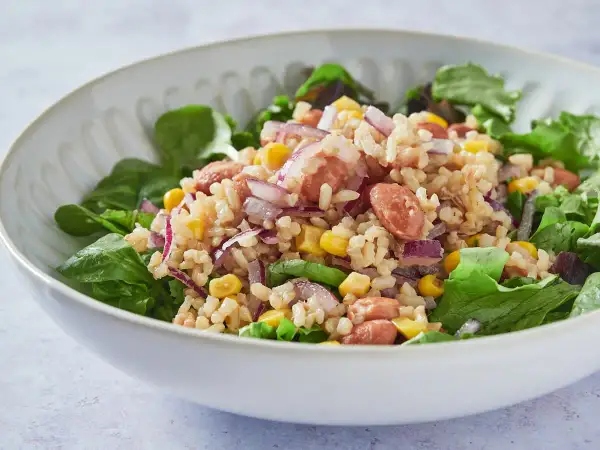 Ensalada de arroz con salchicha, maíz, cebolla morada y frijoles, servida sobre hojas verdes frescas.