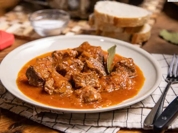 Sabrosa carne con tomate, guisada lentamente hasta quedar tierna. Servida en un plato blanco con pan y cubiertos.