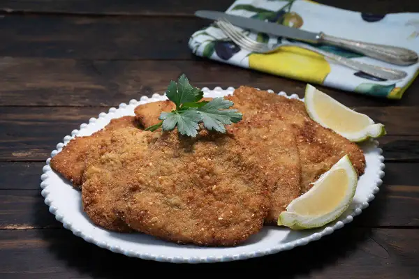 Milanesa de ternera dorada y crujiente, servida en plato blanco con gajos de limón y perejil.